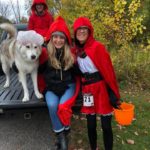 Two women, each dressed as Little Red Riding Hood, pose with their dog. The dog wears a silly hat.