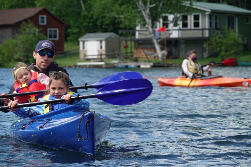 Adaptive kayakers. In the foreground, a man kayaks with two young children. In the background, a kayaker wears a mask
