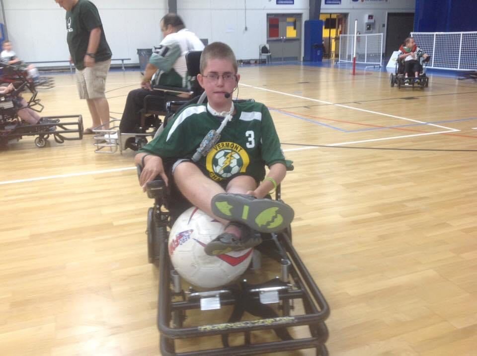 Kyle in a power soccer chair, dribbling the ball with legs outstretched.